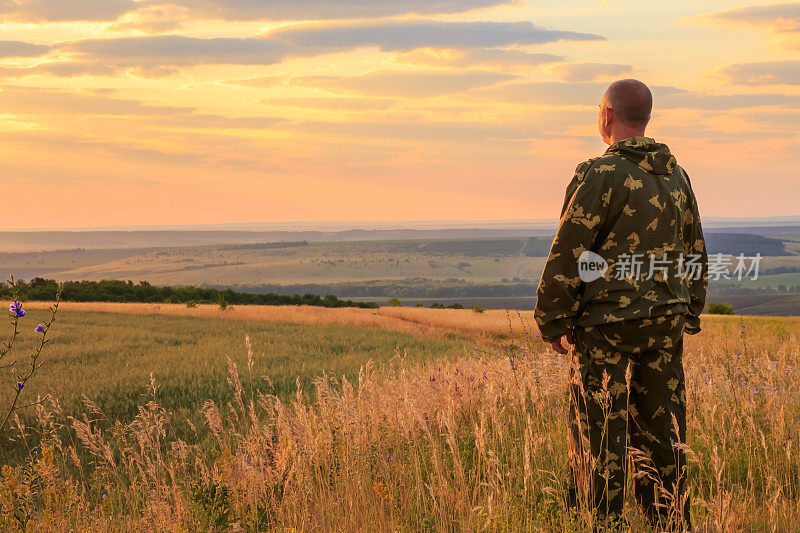 一个40 ~ 45岁的欧洲男性农民，在夏天的黎明，在温暖的阳光下，在夕阳的天空和地平线的背景下，茁壮地站在田地里。眺望远方的人